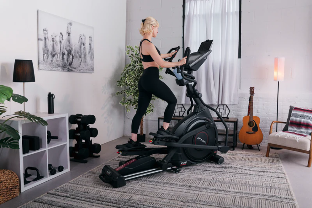 Blonde woman in a black sports bra and black leggings working out on a SOLE E35 elliptical trainer with a mounted display.