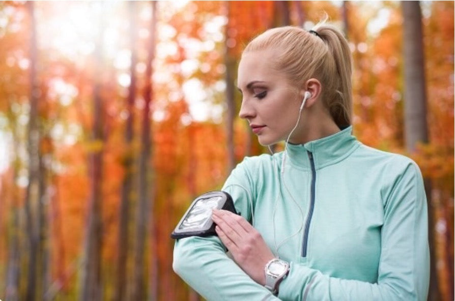 Blonde woman in a teal jacket adjusting a phone armband while wearing earphones among autumn trees