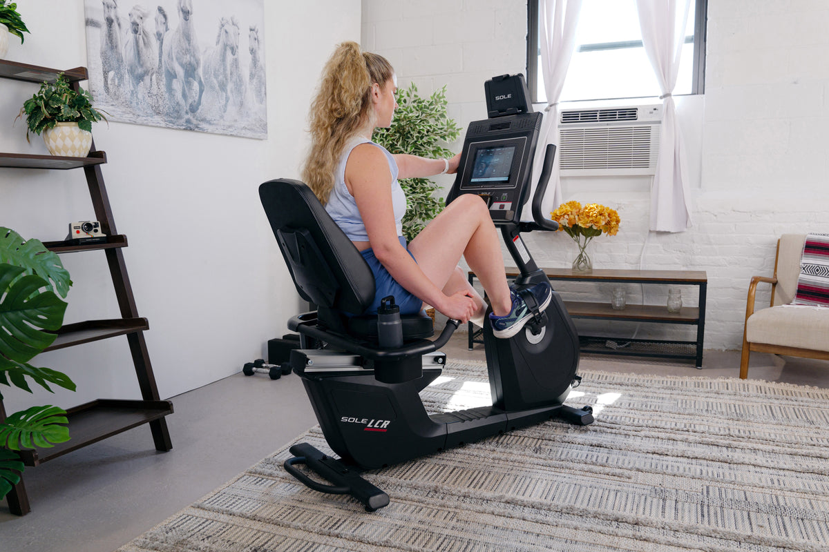 Woman using the SOLE LCR recumbent exercise bike at home, seated in the wide lumbar-supported seat while adjusting the touchscreen.