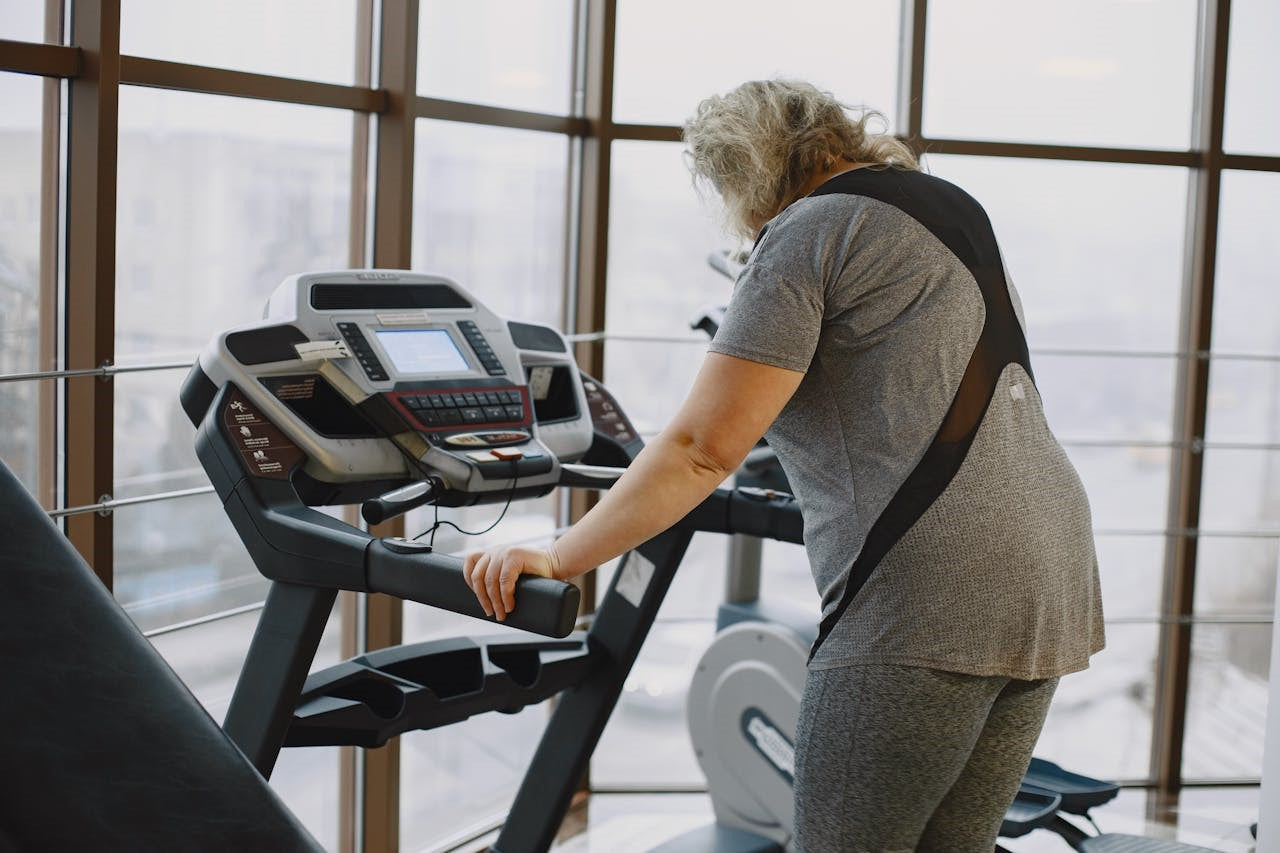 Older woman walking on a treadmill in a well-lit gym with large windows.