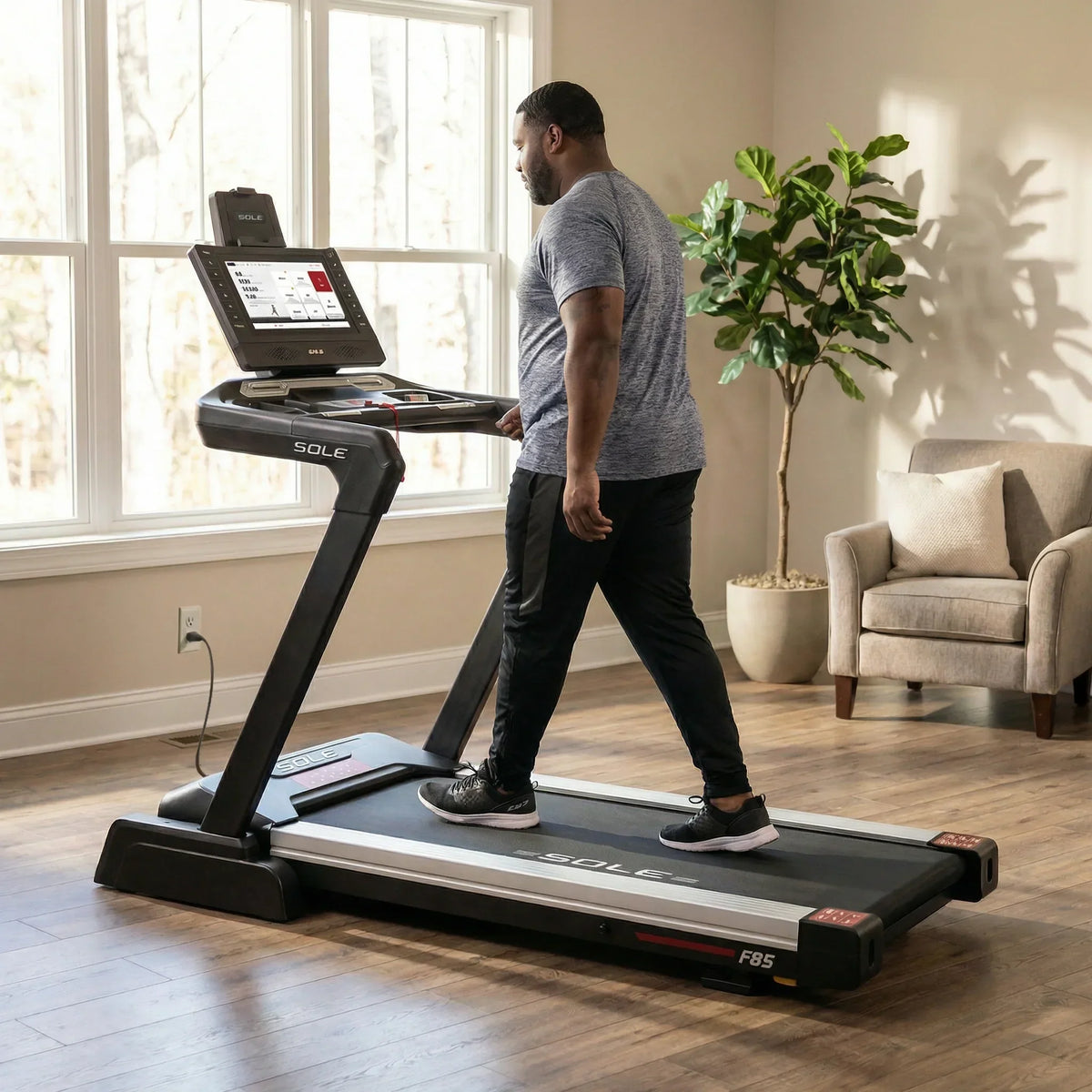 Man with short dark hair and a beard in a gray t-shirt and black track pants walking on a SOLE F85 treadmill with a touchscreen display
