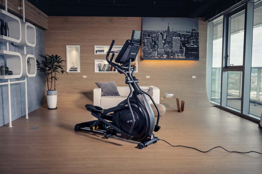 SOLE E25 elliptical trainer with a mounted display and dual handlebars shown from a side angle in the background of a modern room.