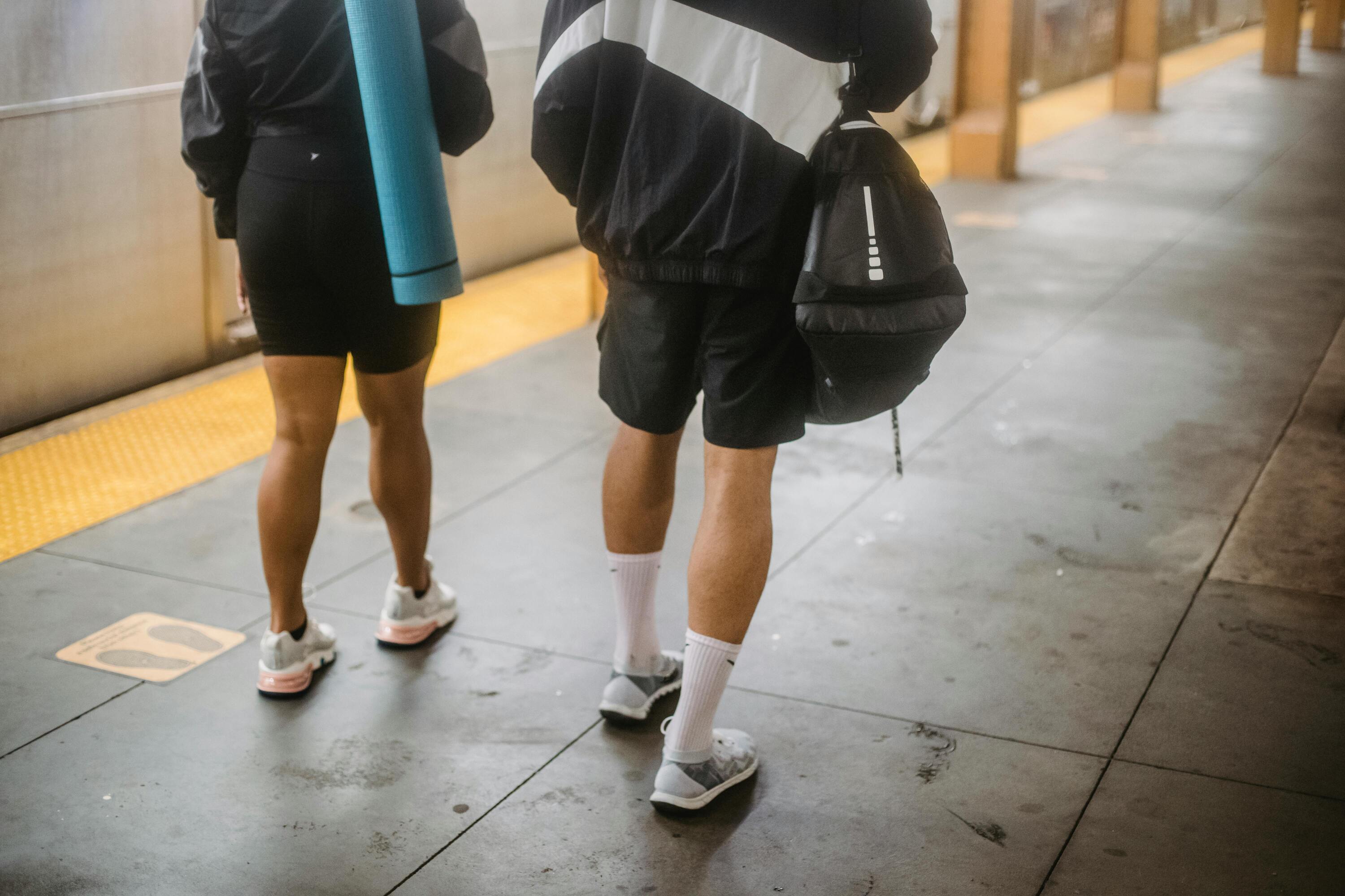 Two people in athletic wear, one carrying a yoga mat and the other a gym bag, standing on a subway platform.