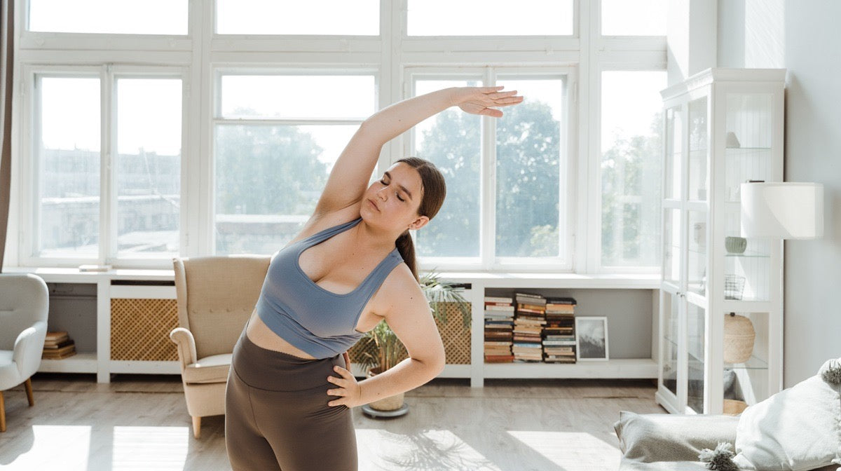Woman doing a standing side bend at home, one arm reaching overhead as she leans to the side.