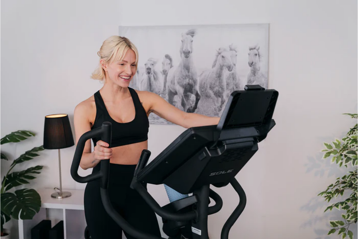 Smiling woman in black activewear using a SOLE elliptical machine in a home gym, showing that low-impact cardio can be enjoyable