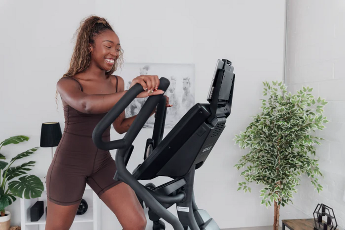 Woman smiling while striding on a Sole elliptical in a bright home gym, showing comfortable low-impact cardio for back pain relief.