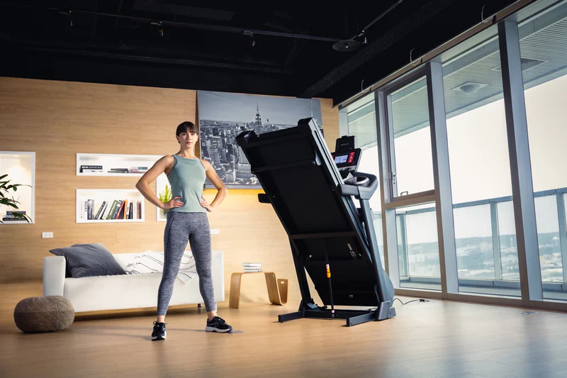 Woman standing confidently beside folded SOLE F63 treadmill in modern, spacious home gym