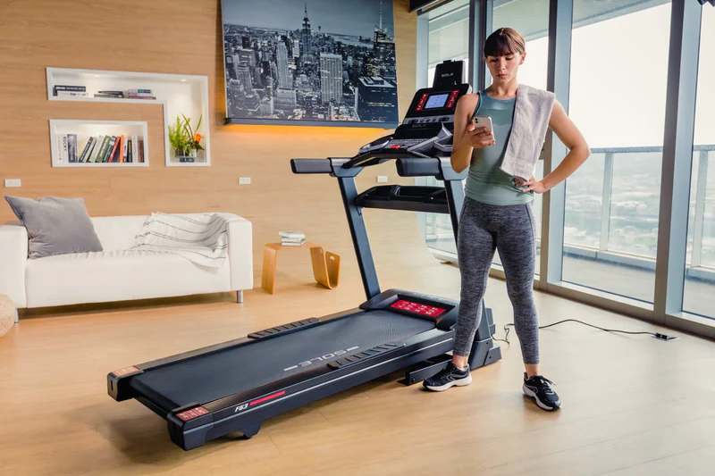 Woman standing next to SOLE F63 treadmill in modern living room, checking phone after workout session.
