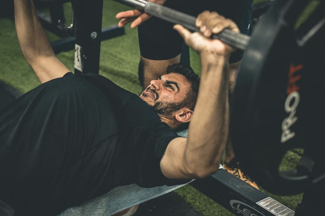 A man in a black t-shirt performs a bench press on an outdoor gym setup, grimacing with effort as he pushes a loaded barbell upward