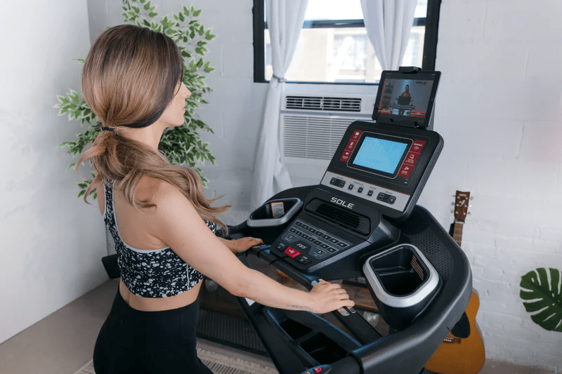 A woman using a SOLE F65 treadmill, watching a workout video on a tablet in a bright, home workout space.