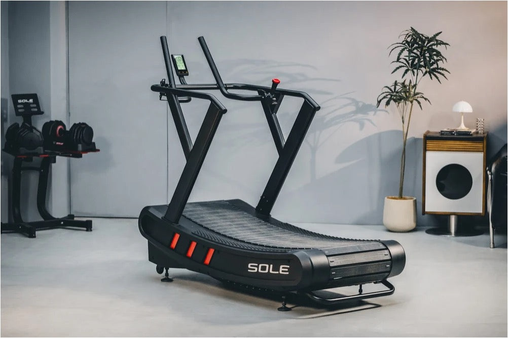 Black Sole curved manual treadmill with red accent lights in a home gym beside a dumbbell rack and a potted plant
