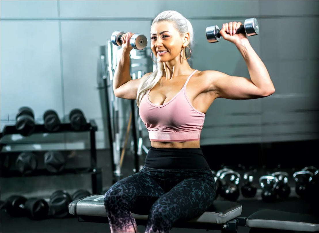 Fit woman performing seated dumbbell shoulder raises in a gym, representing the upper-body strength training central to Camila Mendes' Teela prep
