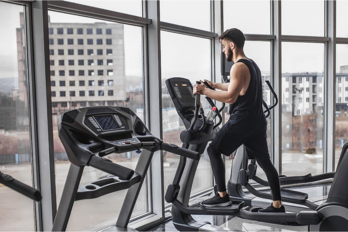 Man in black athletic wear training on an elliptical machine next to a large gym window