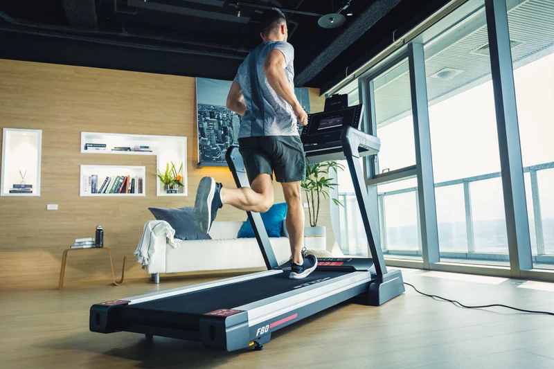 Man running on the SOLE F80 treadmill in a modern open-plan living space with a city wall mural, white sofa, bookshelf, and floor-to-ceiling windows in the background.