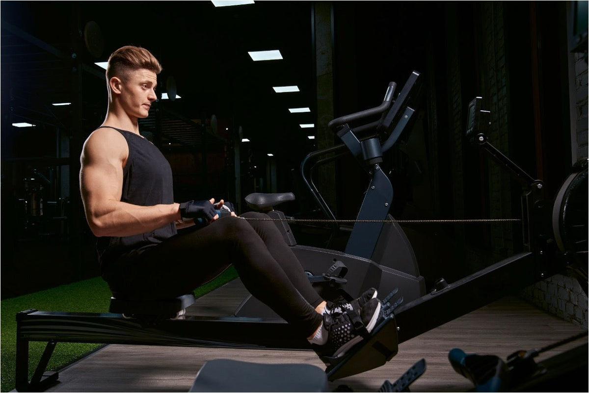 Muscular man in a black tank top rowing on an indoor rowing machine in a dimly lit gym