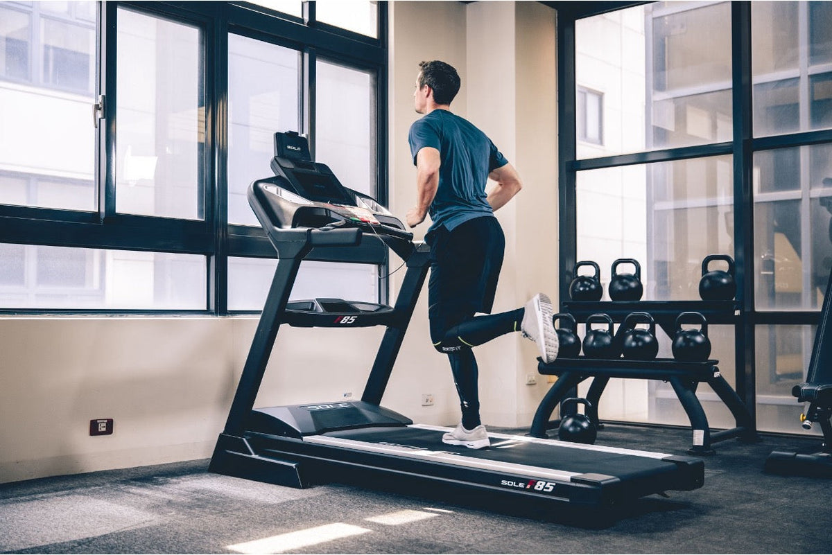 A man in athletic wear runs on a Sole F85 treadmill in a gym beside a rack of kettlebells