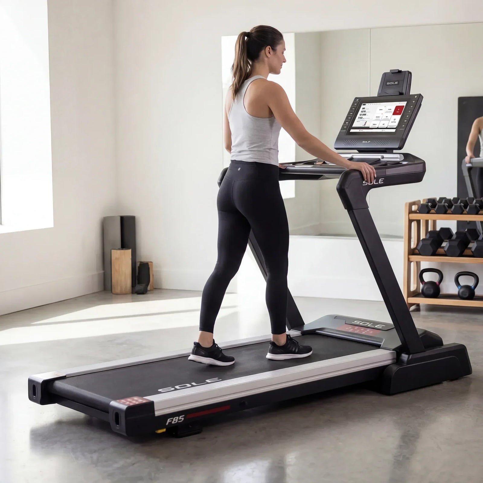 Woman walking on the SOLE F85 treadmill in a minimalist home gym with a large mirror, dumbbell and kettlebell rack, and polished concrete floors in the background.