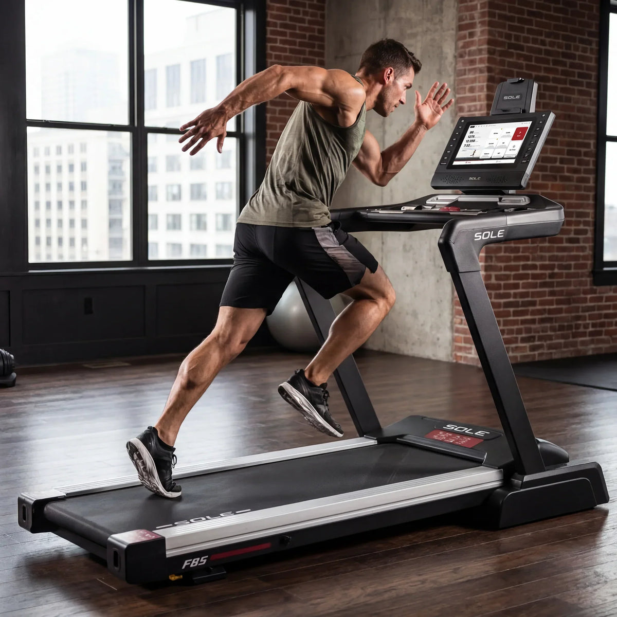 Athletic man with short dark hair in an olive green tank top and black shorts sprinting on a SOLE F85 treadmill with a touchscreen display.