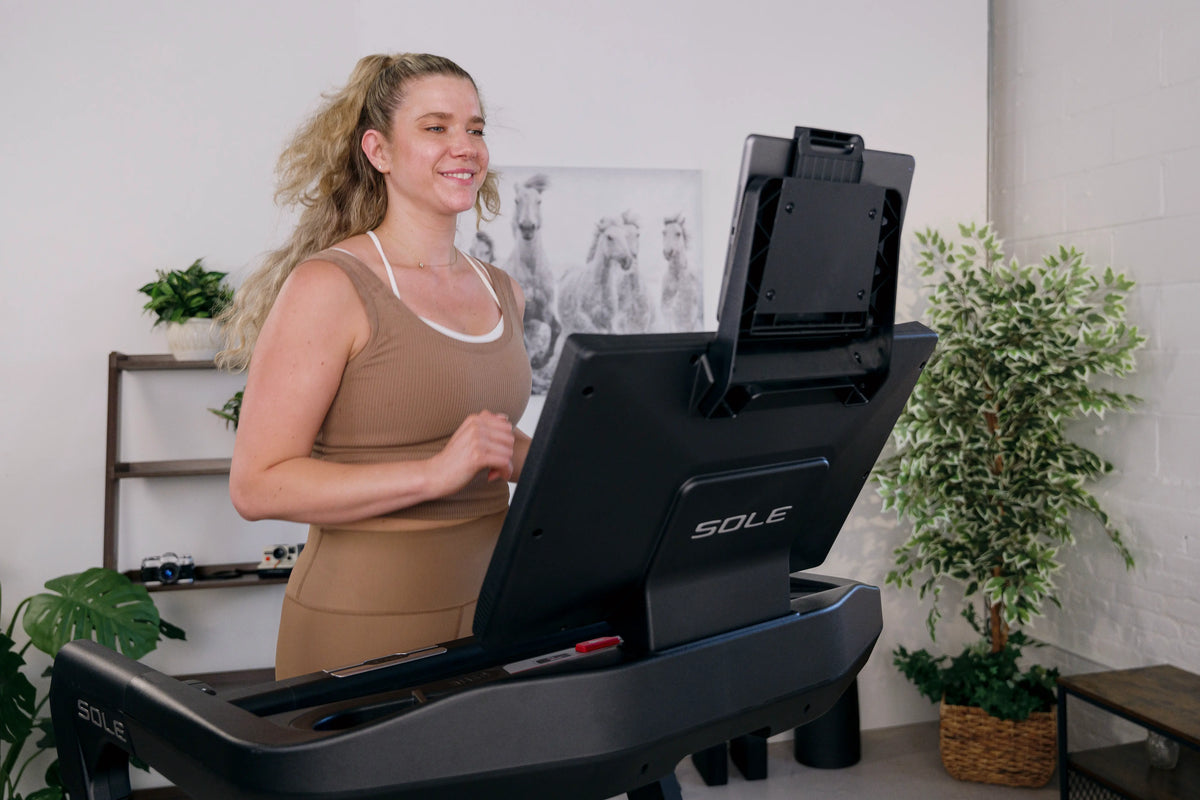 Woman smiling mid-workout on a black SOLE treadmill, showing how it fits naturally into a bright home gym setup.