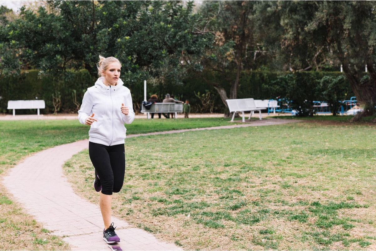 Woman in a white hoodie and black leggings jogging along a paved path through a green park