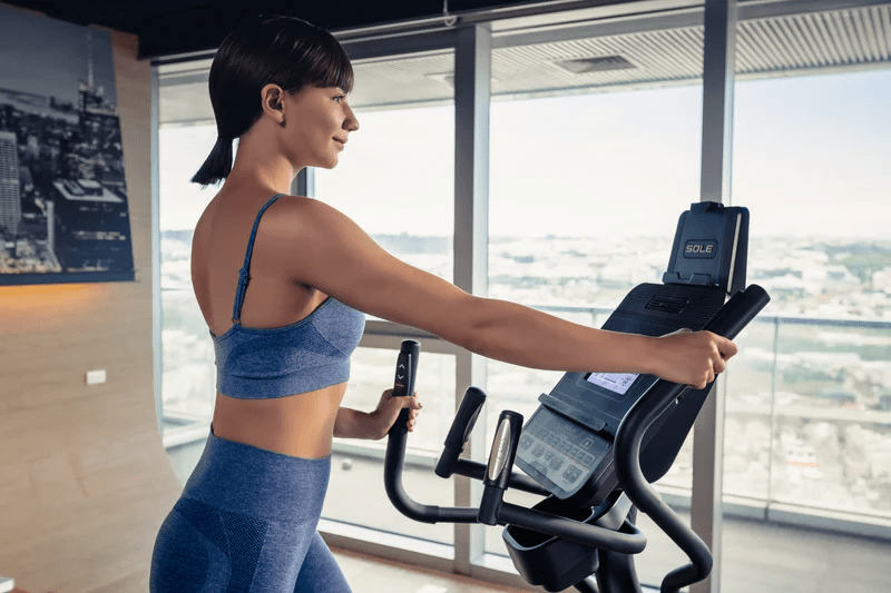 Woman in blue workout outfit uses SOLE elliptical machine in gym with large windows showing cityscape view
