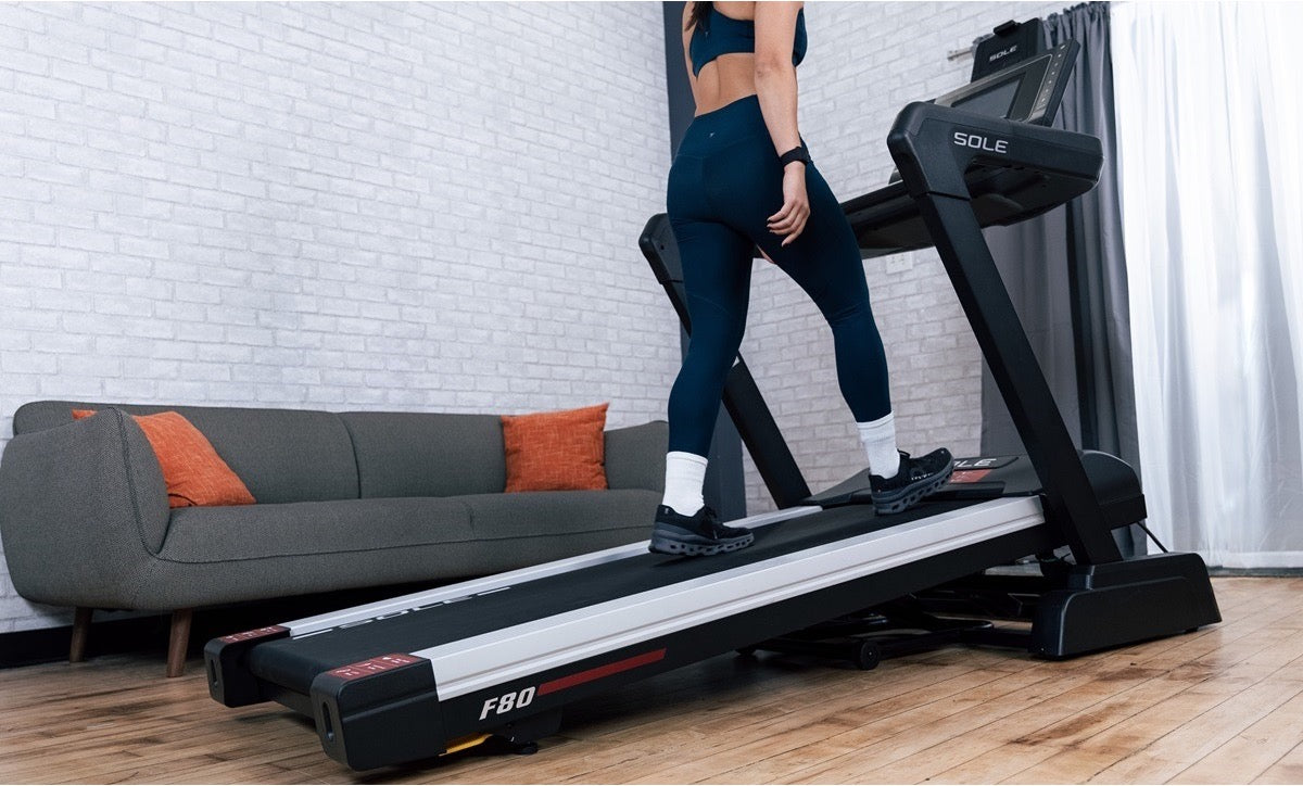 Woman walking on an inclined SOLE F80 treadmill in a living room, with the deck raised to simulate an uphill walk