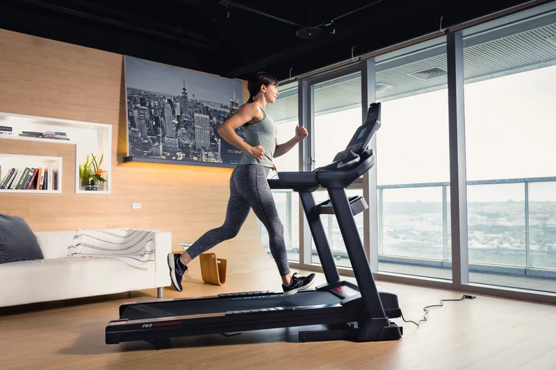Woman wearing sportswear jogging on SOLE treadmill in modern apartment with large windows.