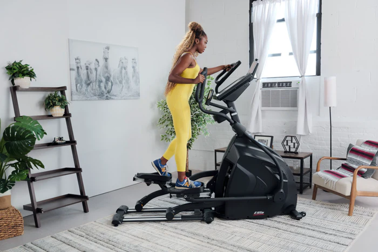 Woman in yellow activewear striding on the SOLE E95S elliptical in a bright, modern home living room.