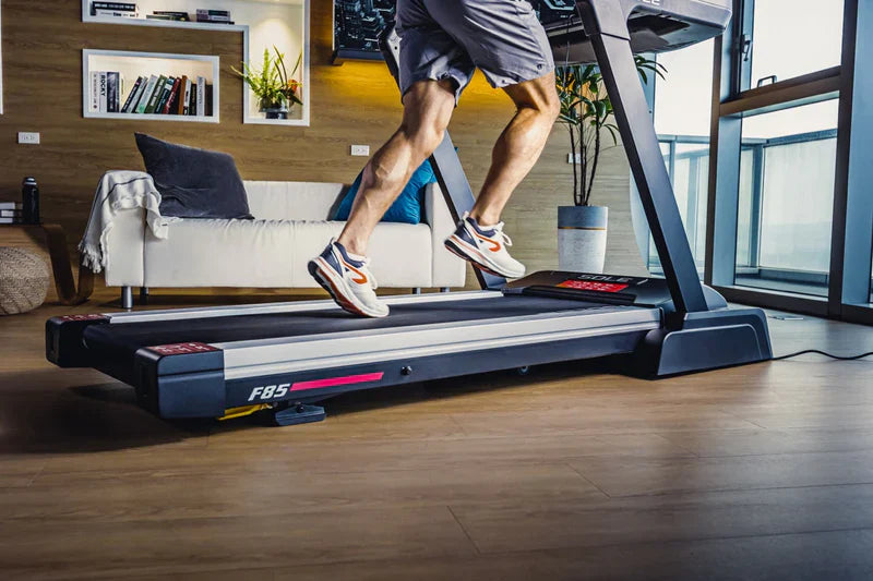 Close-up of a man's legs in gray shorts and white and orange sneakers running on the running deck of a SOLE F85 treadmill.
