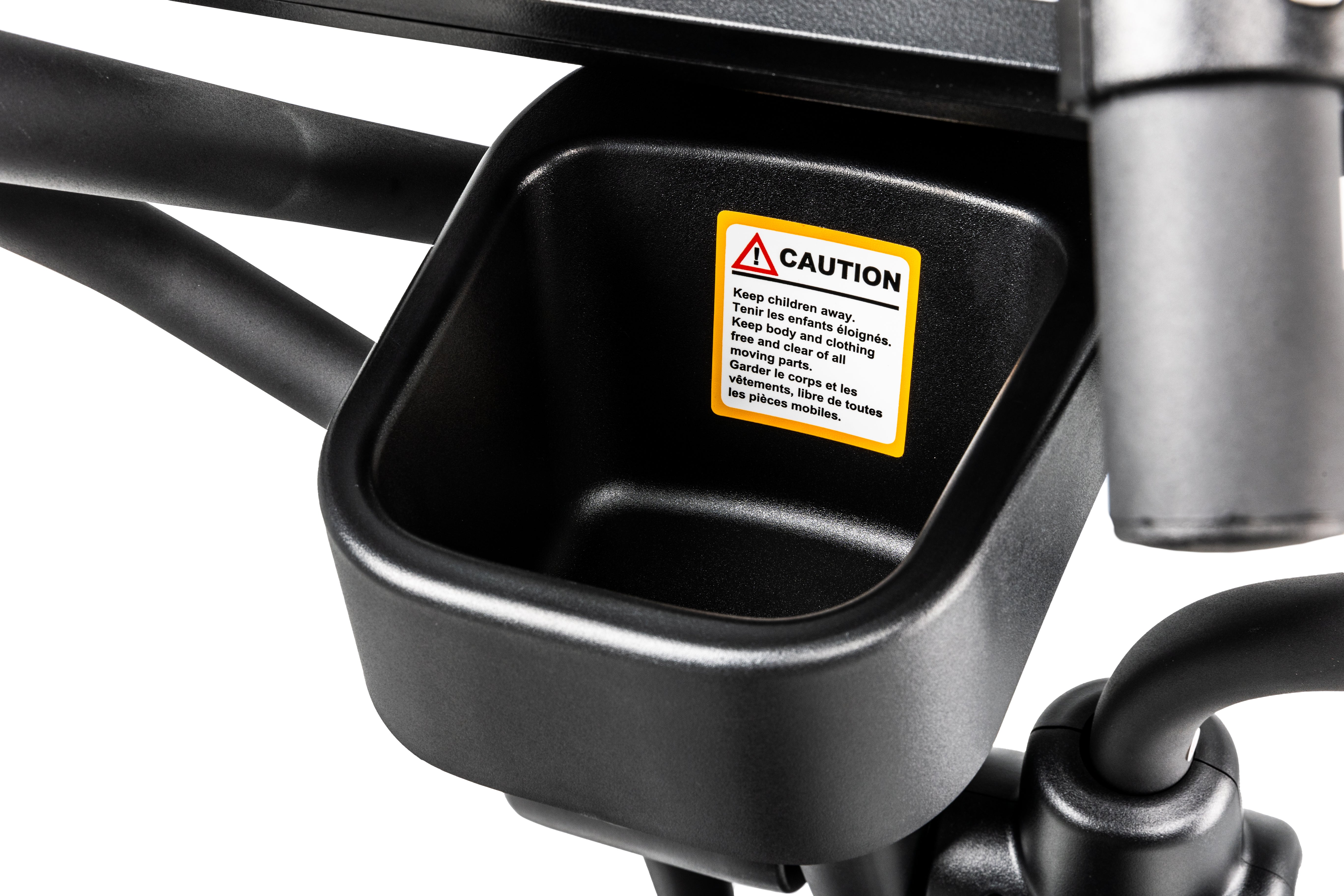 Close-up view of a storage compartment on the Sole E25 elliptical trainer, showing a black tray and surrounding structure, with a prominent yellow 'CAUTION' label warning users to keep children away and be mindful of moving parts.