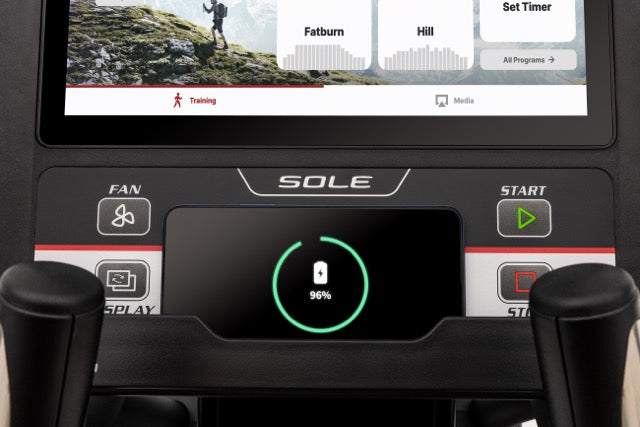 Detailed view of the Sole E98 console displaying a mountain training scene, workout programs like "Fatburn" and "Hill", and a battery indicator at 96%. The console has buttons labeled "FAN", "DISPLAY", and "START" alongside the SOLE branding.