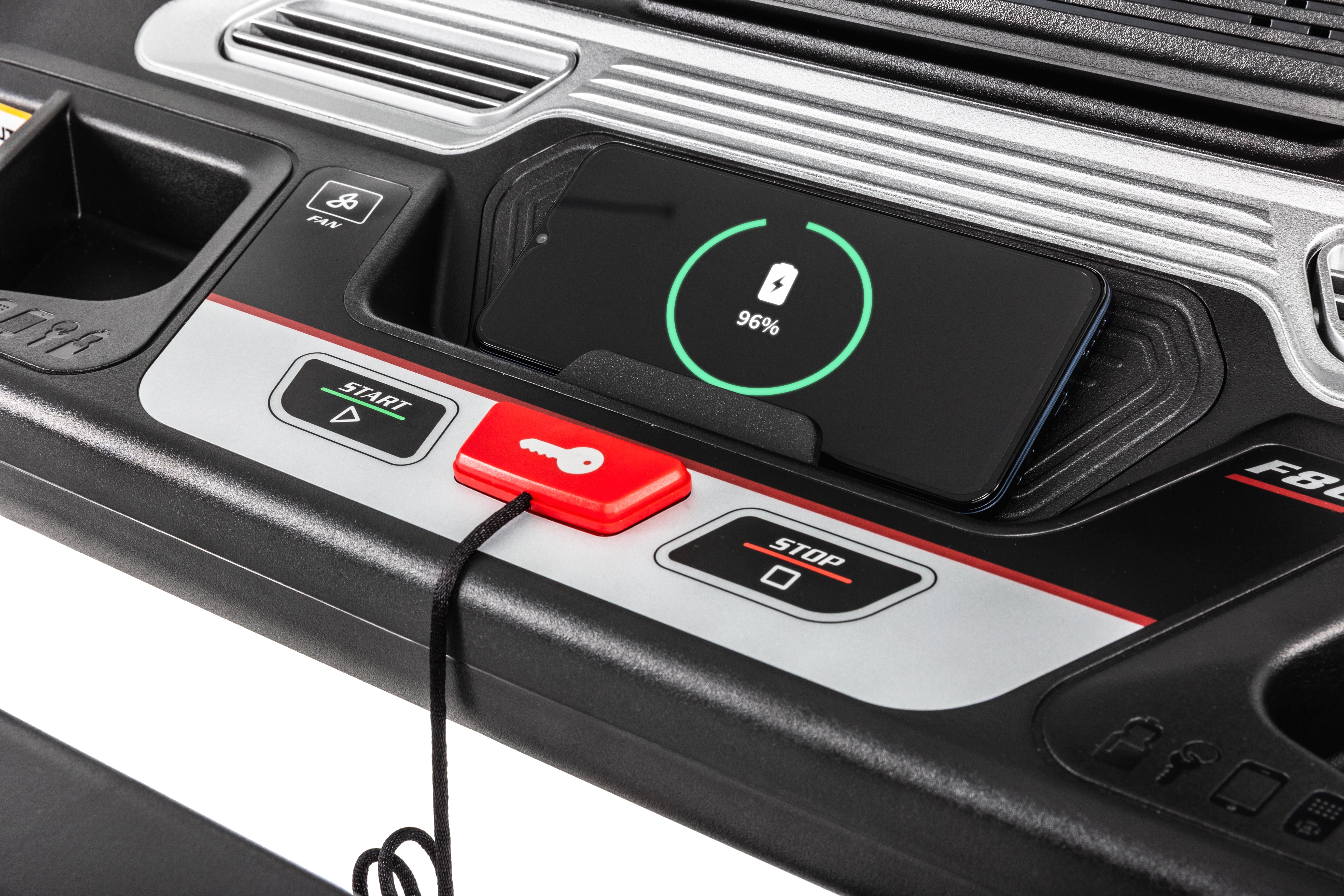 Close-up of the Sole F80 treadmill's control panel showing a smartphone with a 96% charging indicator placed on the device holder. The panel includes a start button, a stop button, speed adjustment controls, a safety key, and an integrated charging port with a connected cable.