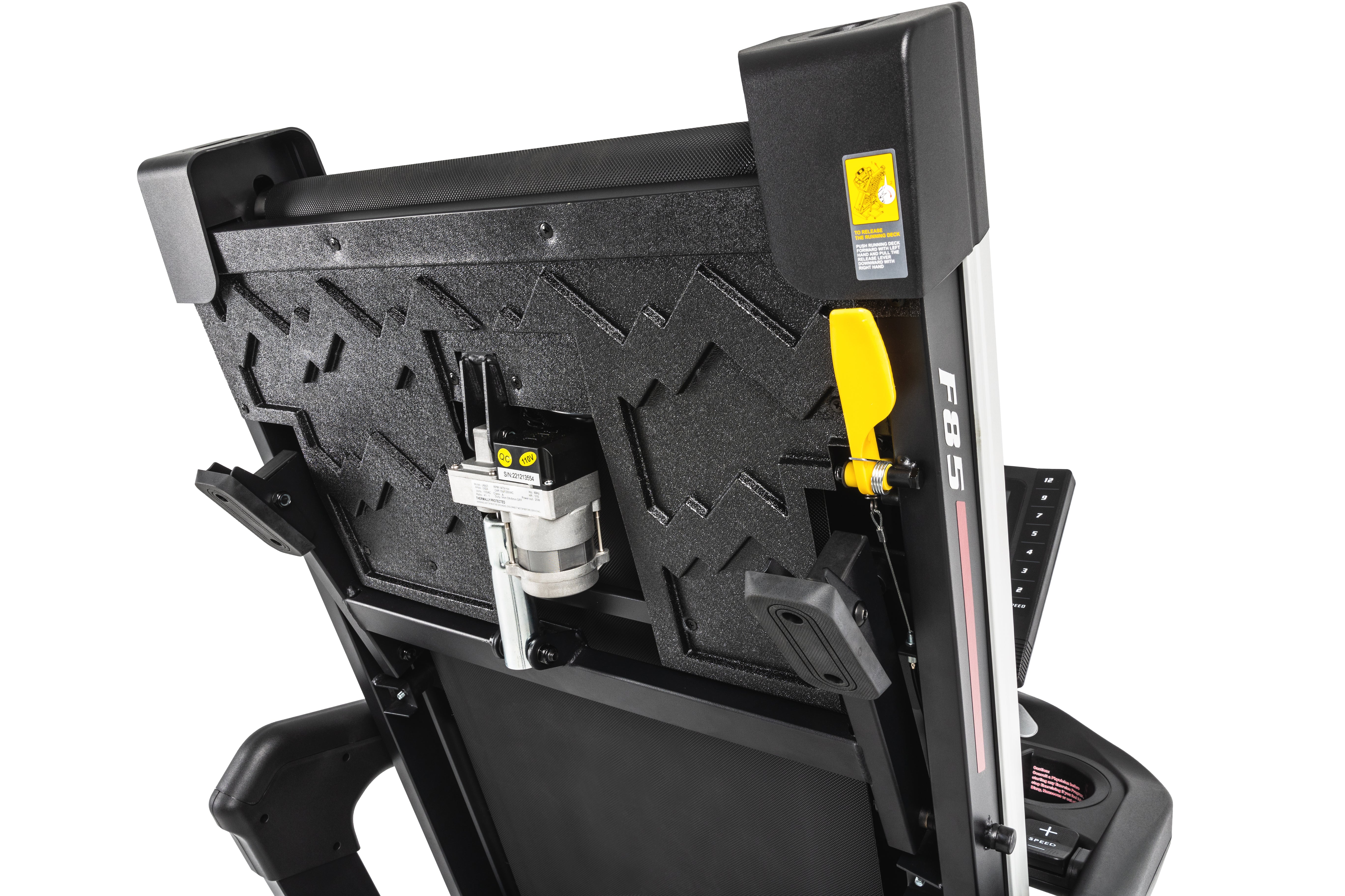 Close-up side view of the Sole F85 treadmill's mechanical components, showcasing a textured deck, a yellow folding release lever, a warning label, and a section of the F85 branding on the treadmill's frame, with a glimpse of the control panel in the background.